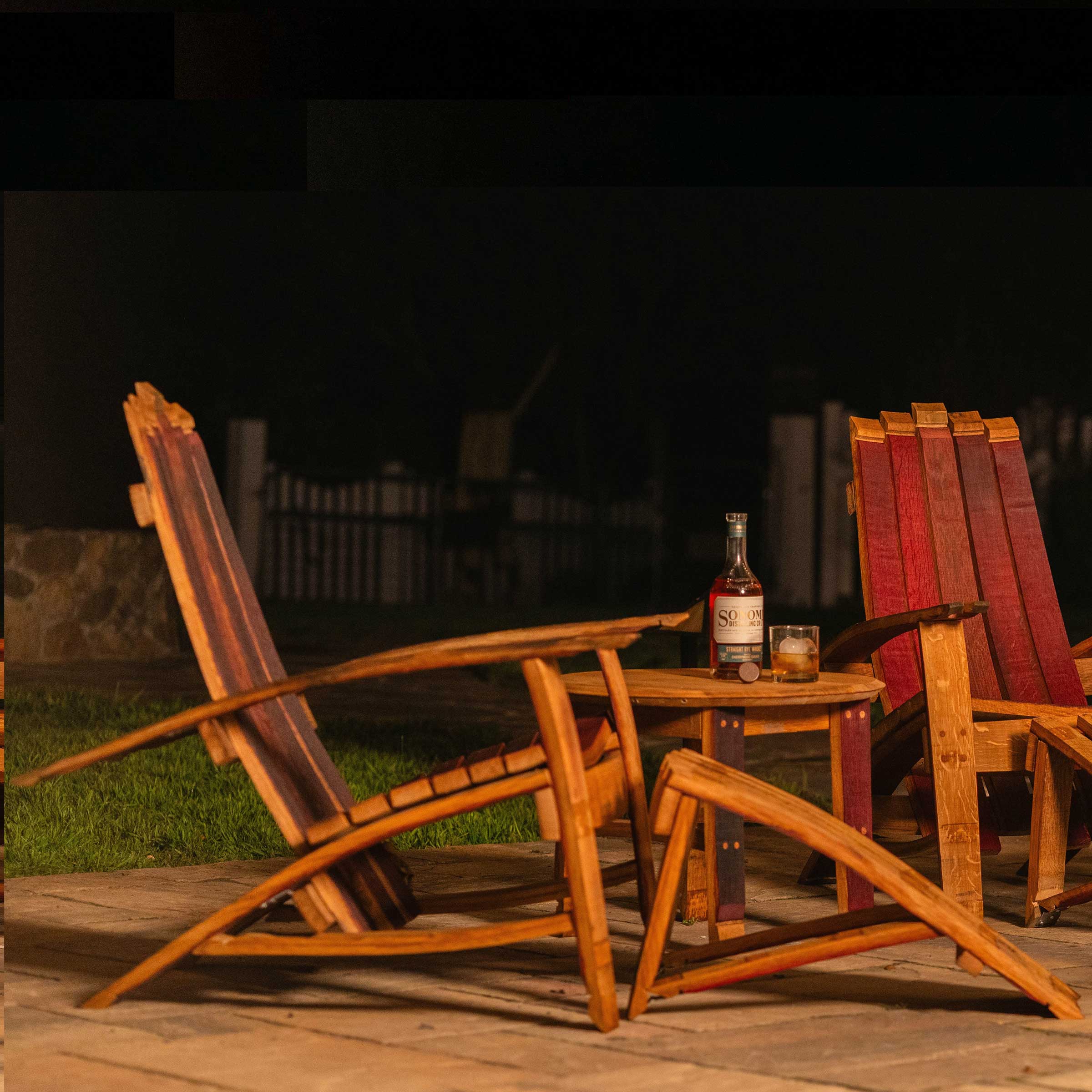 Two Cellr wooden Adirondack chairs with matching footstool and side table made from reclaimed wine barrels, shown outdoors at night with a bottle of wine on the table.