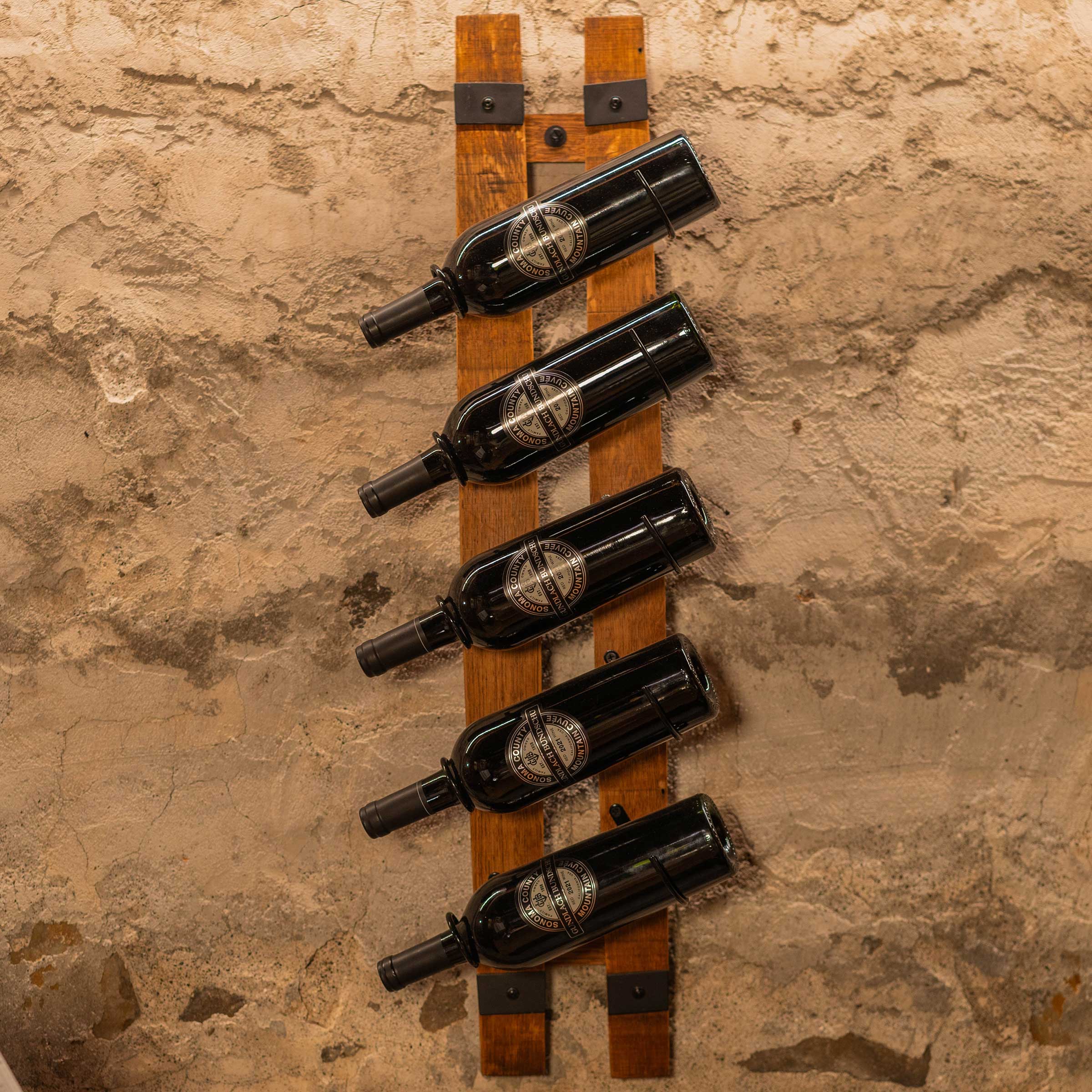 Five wine bottles arranged vertically on two parallel reclaimed barrel staves mounted to a textured wall.