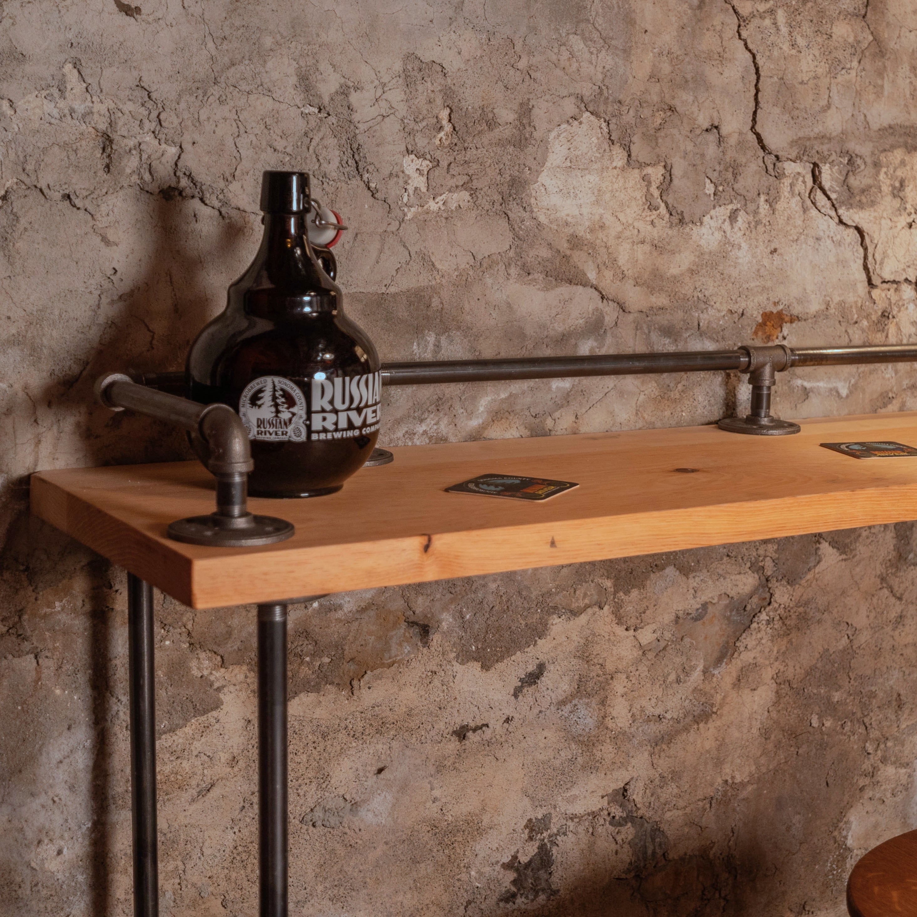 Close-up of the Planq High Top Bar Table showing the reclaimed wood top and black iron pipe flange mount against a stone wall.”