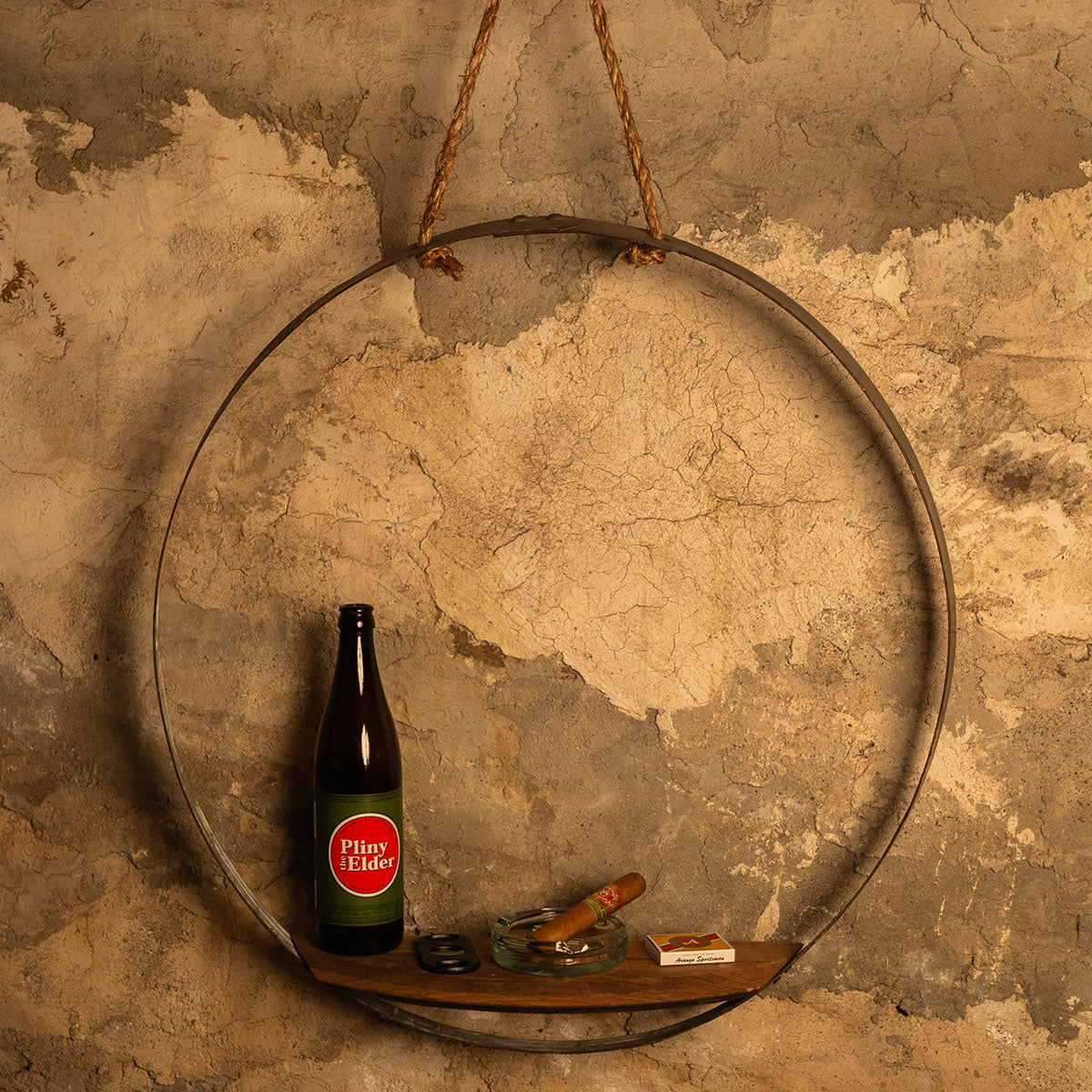 Rustic round wall shelf made from a reclaimed wine-barrel hoop with a wooden half-moon shelf, hanging by rope on a textured stone wall.
