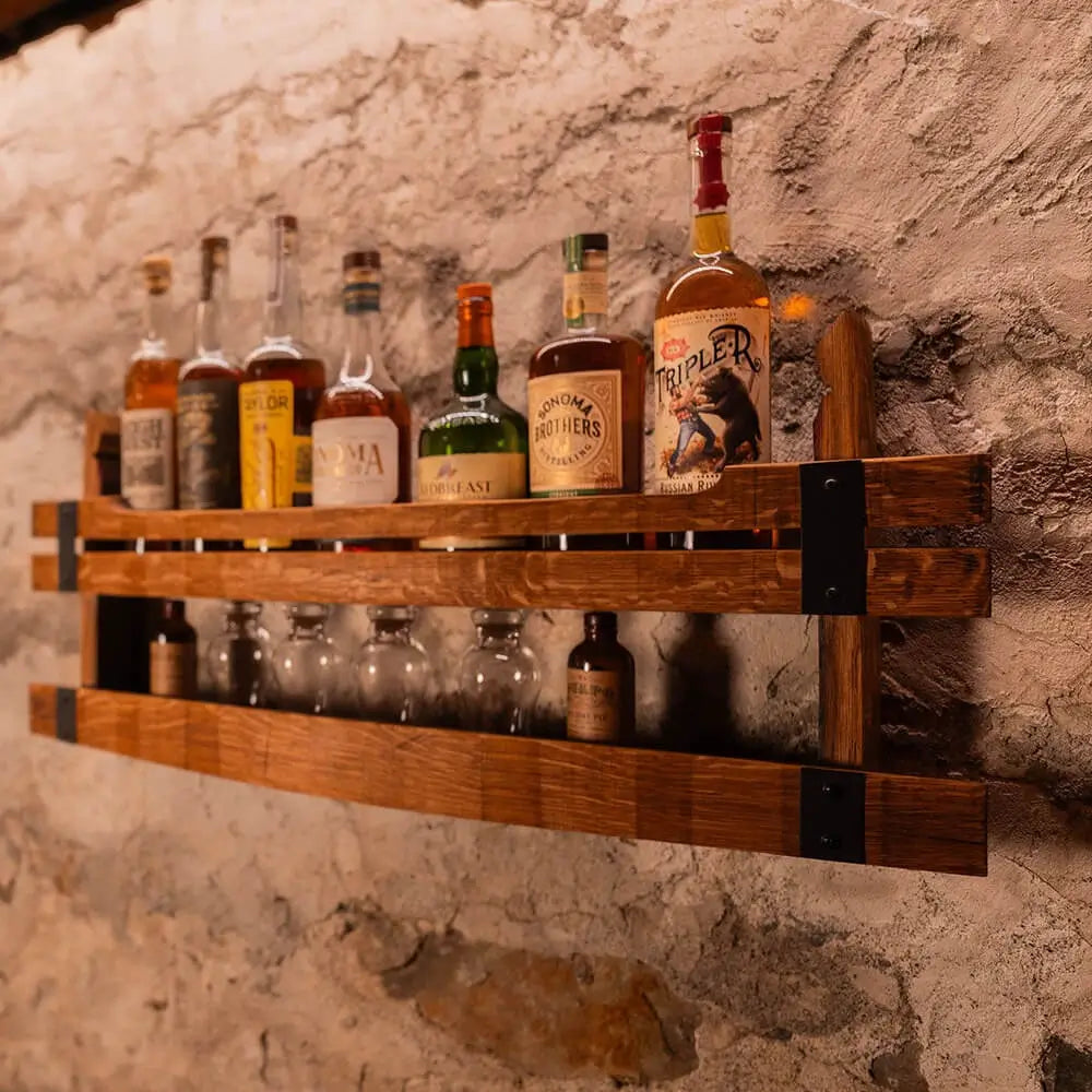 Two-tier wooden wall shelf holding liquor bottles on the top shelf and glasses on the lower shelf against a stone wall.