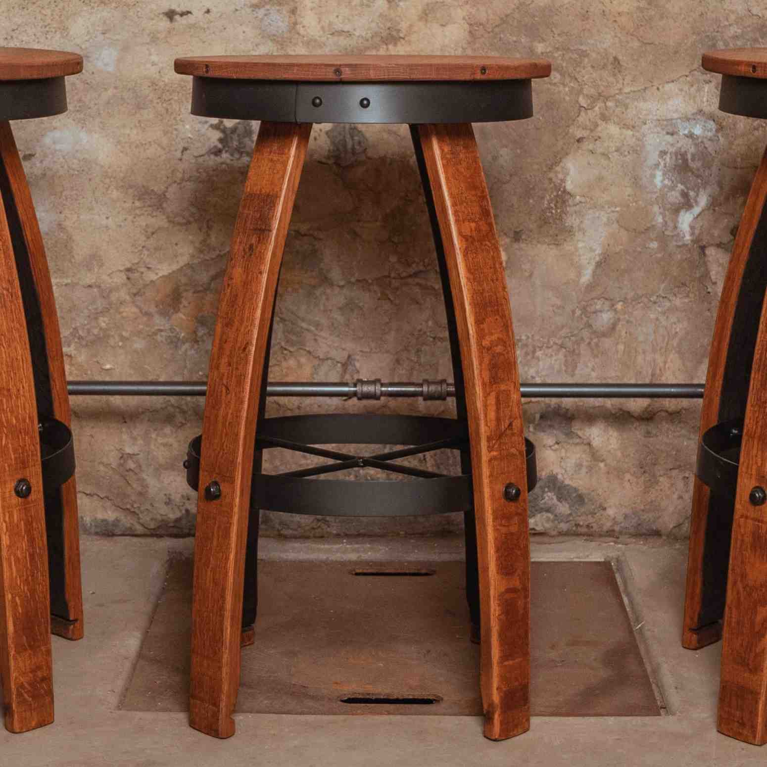Set of three reclaimed wine barrel bar stools with curved oak staves, round wooden seats, and black metal footrest hoops arranged in a rustic cellar-style room.