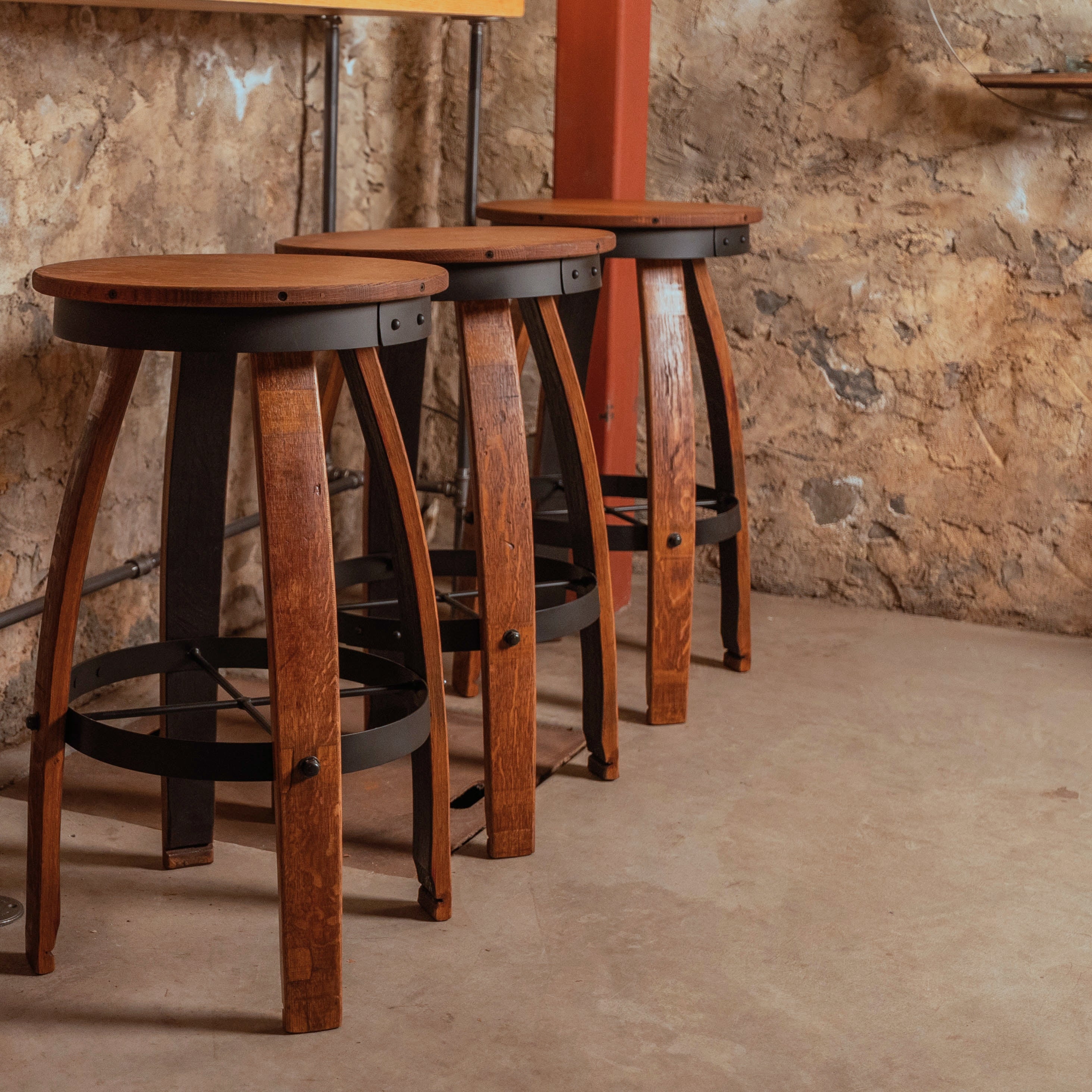 Close-up of a rustic bar stool handcrafted from reclaimed wine barrel staves, featuring a round wooden seat, black metal hoop, and subtle industrial accents against a stone cellar wall.