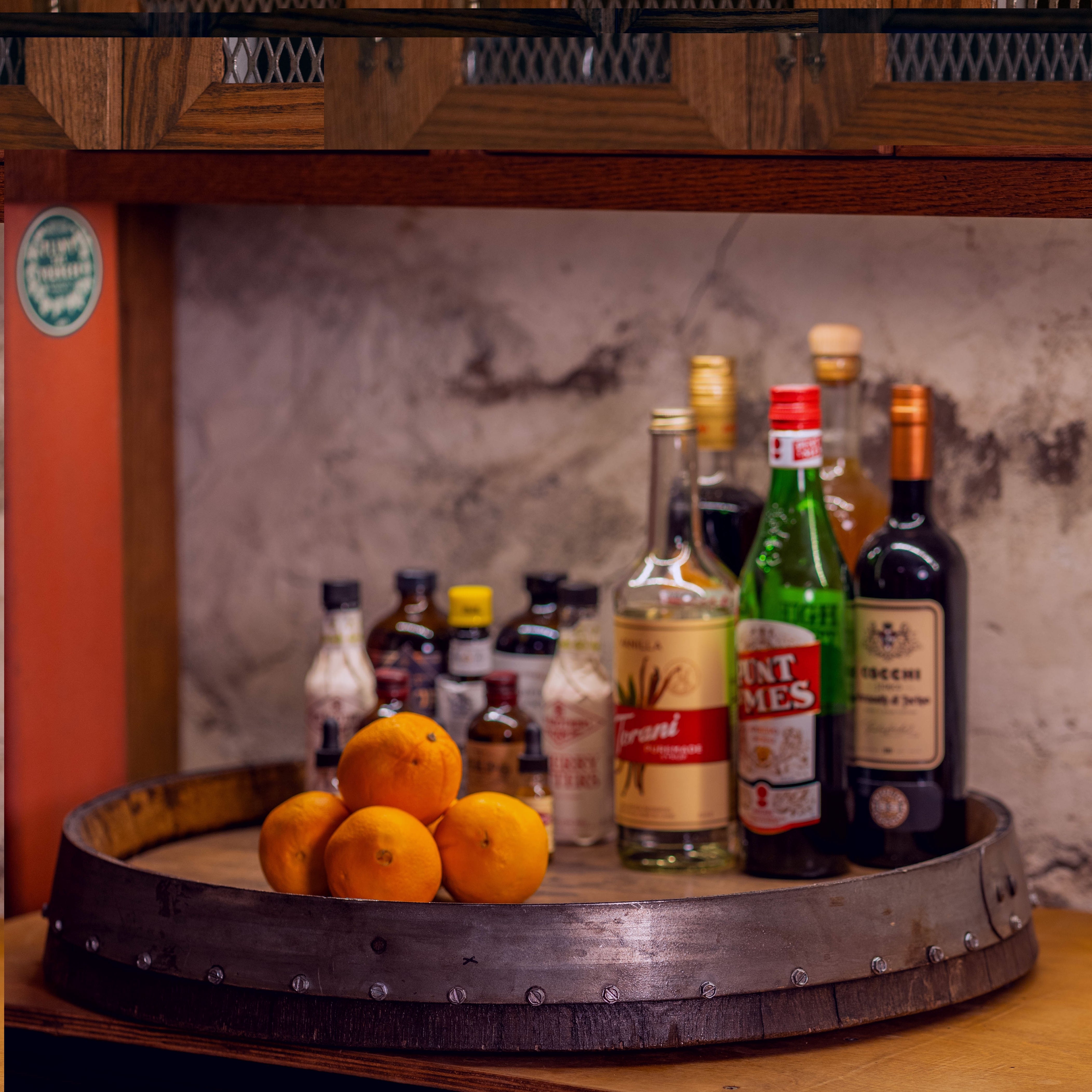 24-inch reclaimed barrel-head Lazy Susan holding liquor bottles, bitters, and oranges on a countertop.