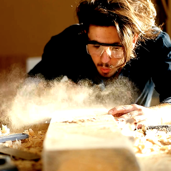 Artisan blowing sawdust from a board, capturing the production process of building reclaimed wood furniture.