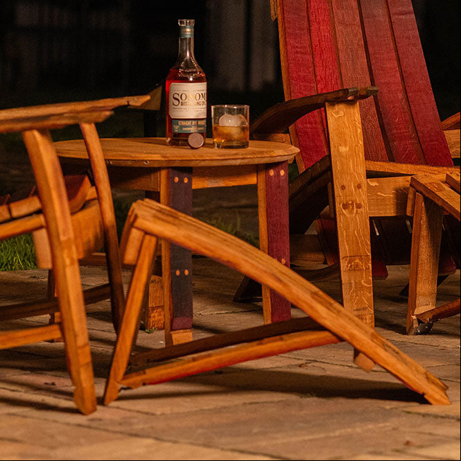 Two Cellr wooden Adirondack chairs with matching footstool and side table made from reclaimed wine barrels, shown outdoors at night with a bottle of wine on the table.