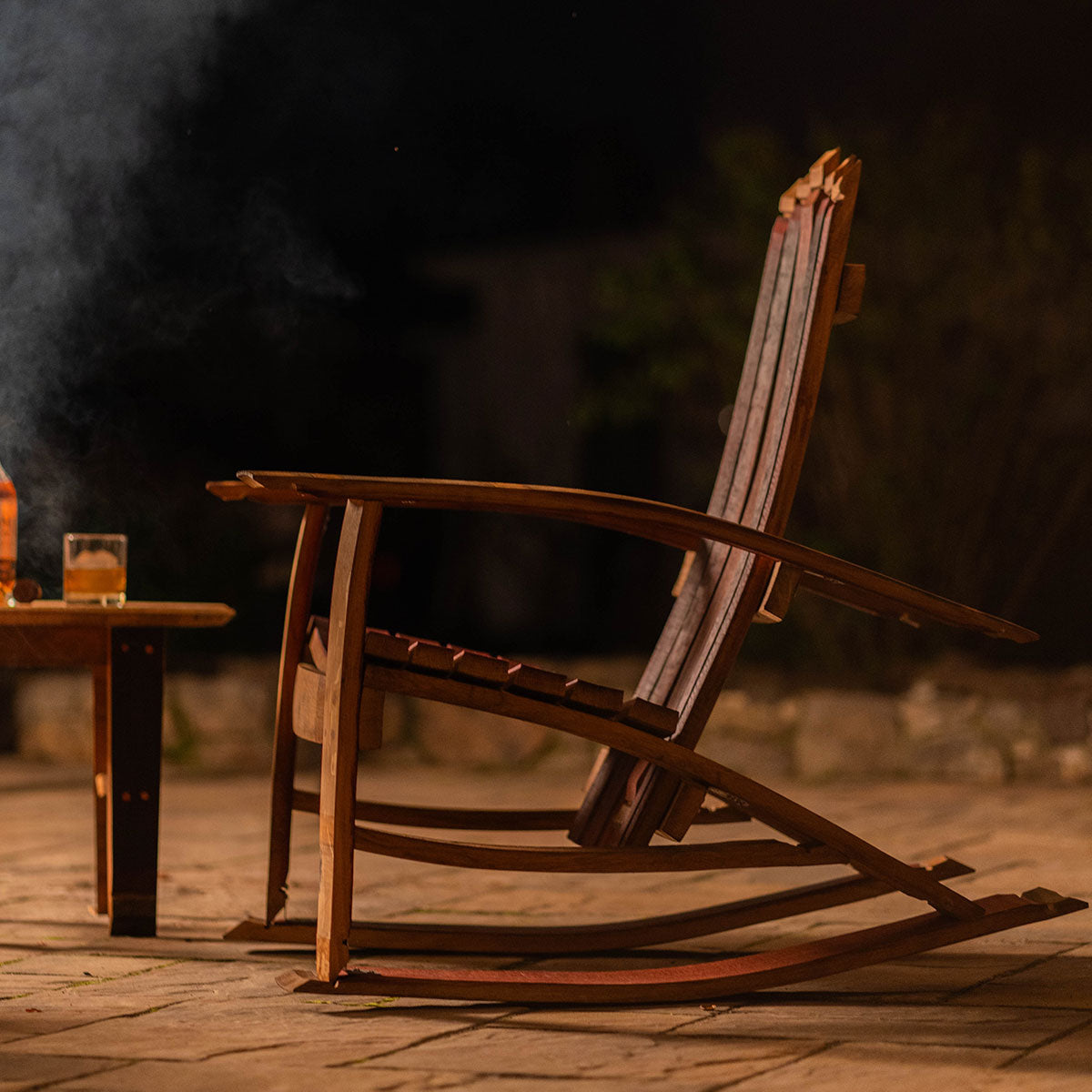 Side view of a handcrafted wooden Adirondack rocking chair made from reclaimed wine barrels, positioned outdoors at night beside a wood table with a drink.
