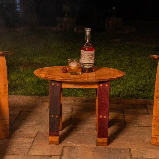 Handcrafted 24" round wood outdoor side table made from reclaimed wine barrel head and staves, displayed on a patio at night with a bottle and glass on top.
