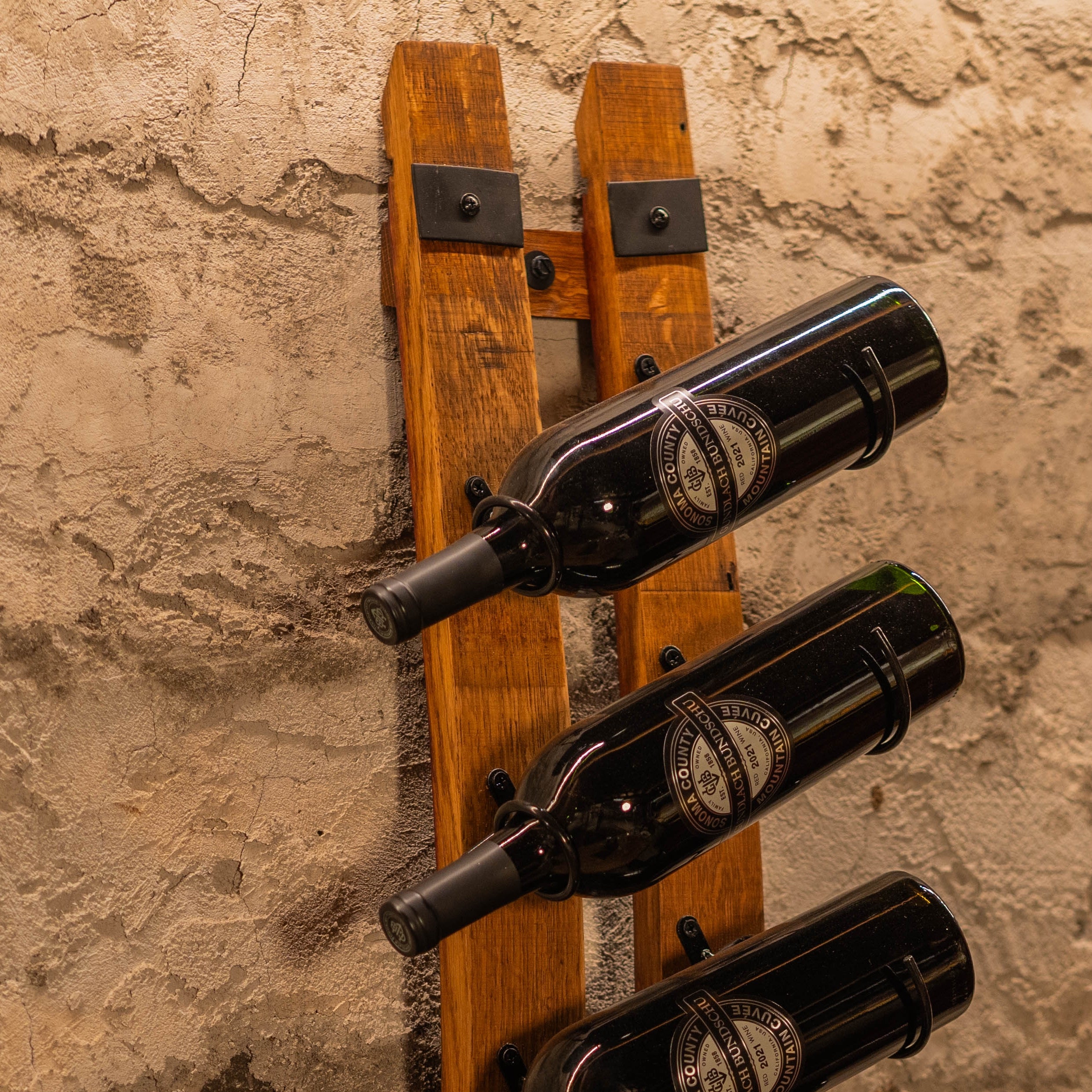 Angled view of a vertical wall-mounted wine rack made from two reclaimed barrel staves holding several bottles.