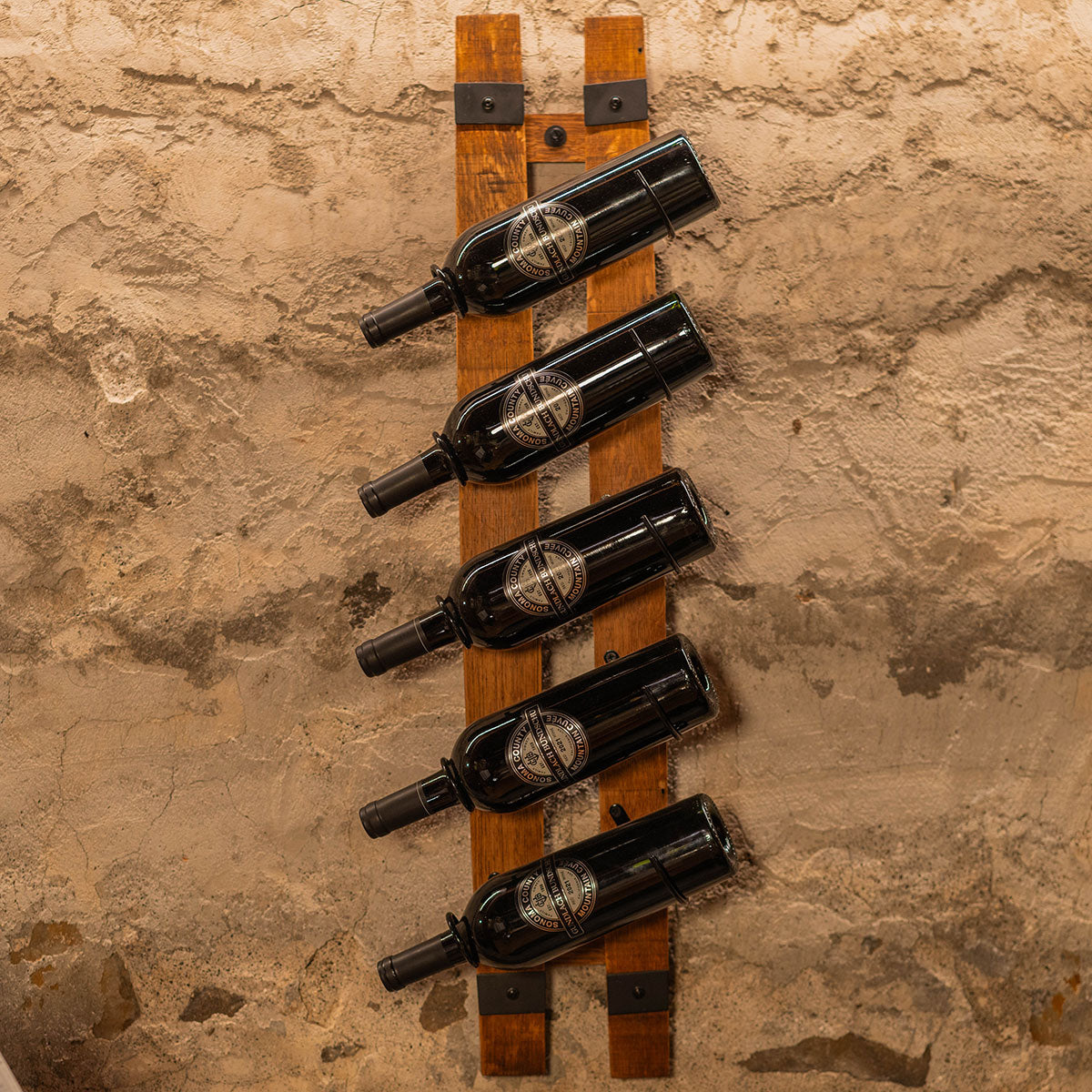 Five wine bottles arranged vertically on two parallel reclaimed barrel staves mounted to a textured wall.