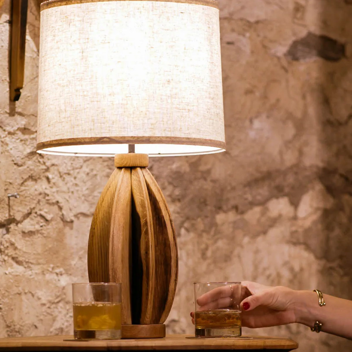 Wooden table lamp with a textured white shade on a stone surface, with a hand holding a glass.