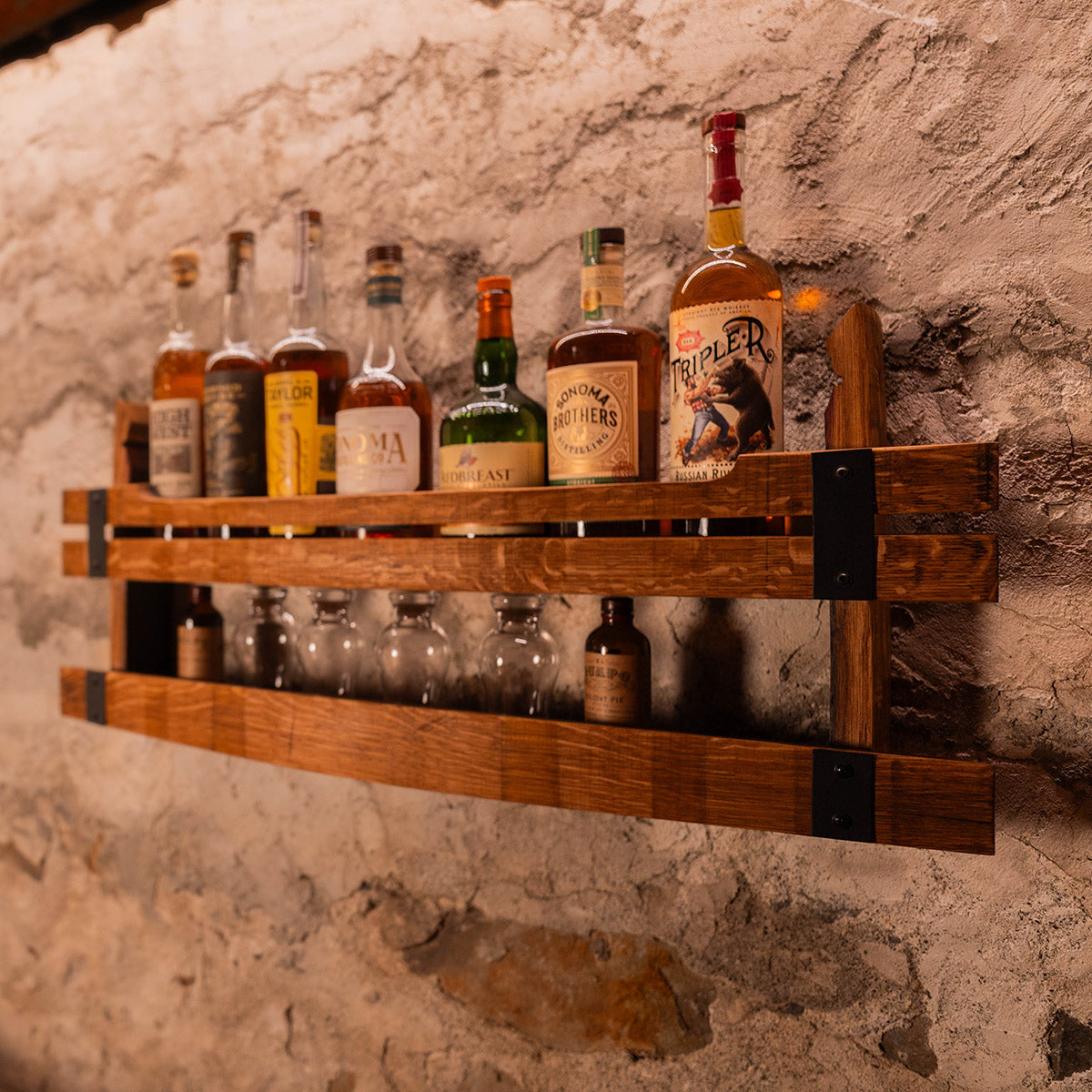 Two-tier wooden wall shelf holding liquor bottles on the top shelf and glasses on the lower shelf against a stone wall.