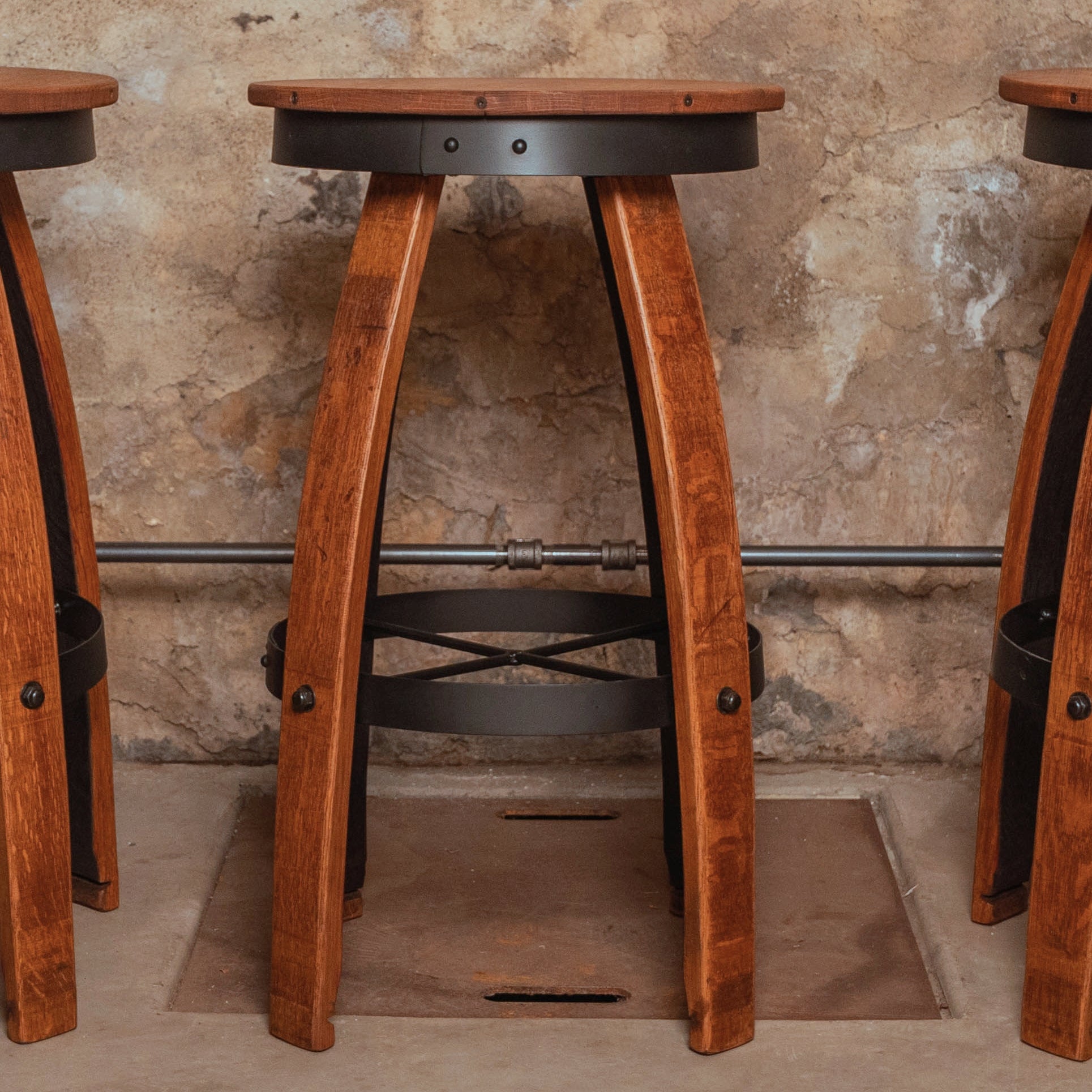 Set of three reclaimed wine barrel bar stools with curved oak staves, round wooden seats, and black metal footrest hoops arranged in a rustic cellar-style room.
