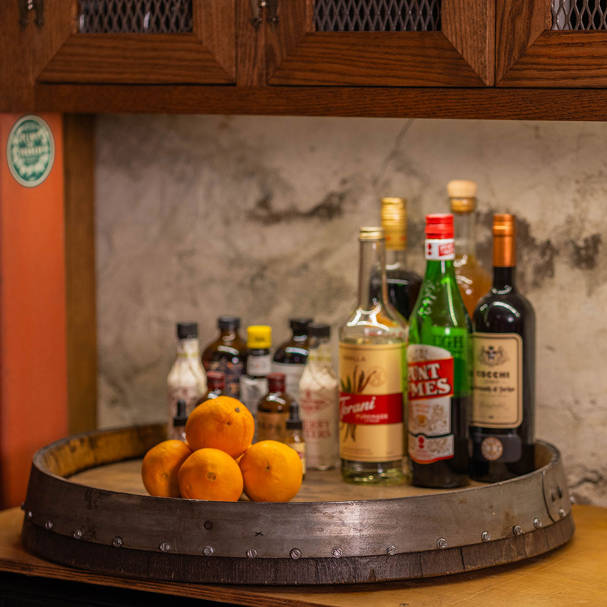 24-inch reclaimed barrel-head Lazy Susan holding liquor bottles, bitters, and oranges on a countertop.
