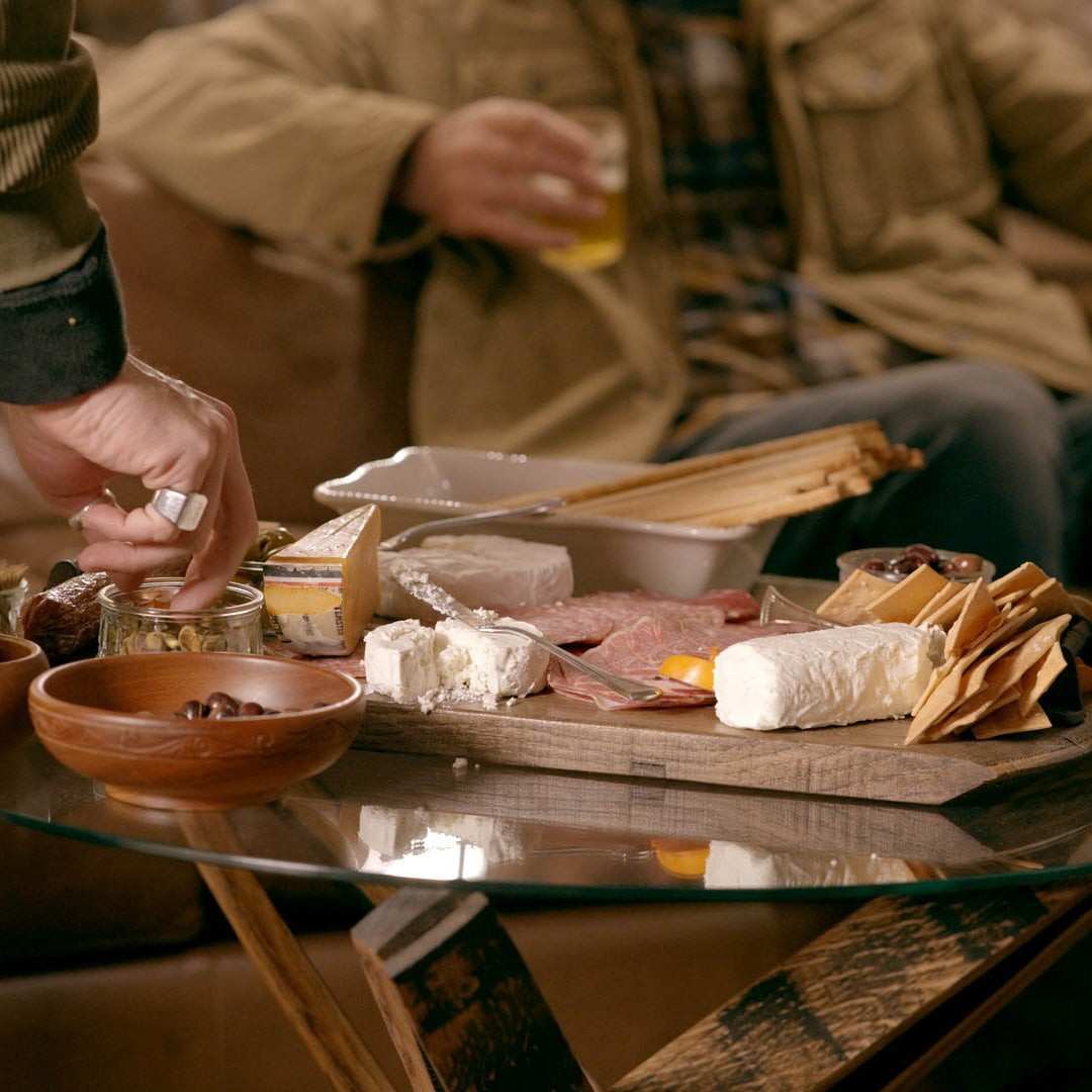 Person preparing a cheeseboard with various items on a glass table, with another person in the background holding a drink.
