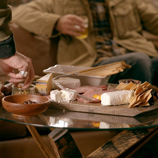 Person preparing a cheeseboard with various items on a glass table, with another person in the background holding a drink.